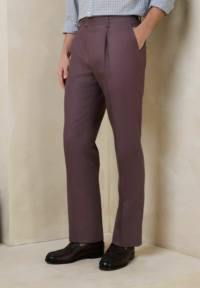 Homme portant un pantalon marron ajusté avec des plis et des mocassins en cuir noir, debout contre un mur beige avec une main dans la poche.