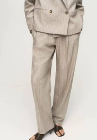 Costume beige clair à rayures avec une veste croisée et un pantalon sur mesure. Tissu lisse avec un léger éclat. Sandales sombres à bout ouvert.