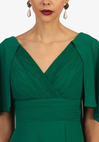 Grünes Kleid mit gefälteltem Oberteil, V-Ausschnitt und fließenden Kapellärmeln. Begleitet von goldenen und Perlen-Ohrringen. Geeignet für formelle Anlässe.