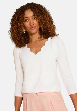 Kobieta o kręconych włosach ubrana w biały top z falistym dekoltem i jasnoróżowe spodnie, zwrócona twarzą w stronę obserwatora na jednolitym tle.