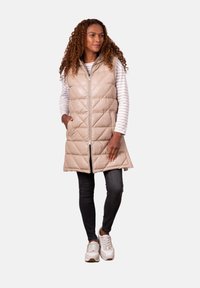 Beige quilted puffer vest with a front zipper, side pockets, and a high collar, worn over a striped long-sleeve top, paired with dark jeans.