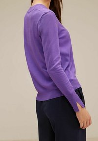 Femme portant un pull violet ajusté à manches longues avec une fente au poignet sur le côté, associé à un pantalon bleu marine foncé, debout devant un fond neutre.