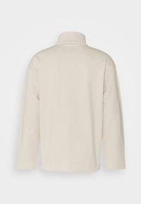 Polaire beige à manches longues avec col montant et coupe décontractée, vue de dos sur un fond clair uni.