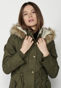 Olive krāsas parka jaka ar mākslīgās kažokādas oderi kapuci, rāvējslēdzēju un pogu aizdari, krūšu kabatu un sašaurināmu jostu viduklī.