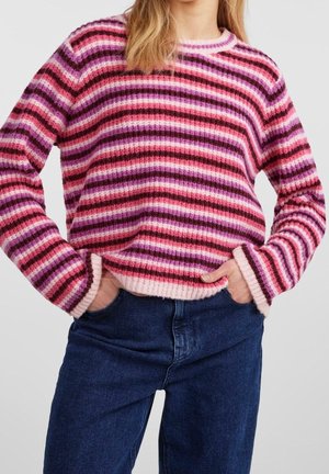 Femme portant un pull en maille rayé rose, violet et rouge avec un jean bleu, les mains dans les poches, devant un fond uni.