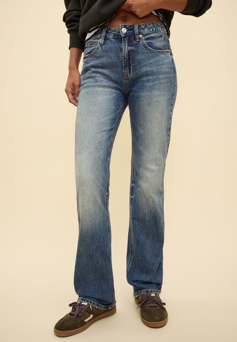 Persona che indossa jeans dritti blu sbiaditi con lacci scuri e scarpe da ginnastica verdi, mano appoggiata sulla vita, sfondo beige.