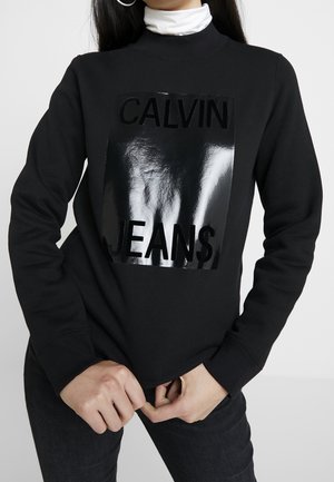Kvinde iført sort sweatshirt med en blank sort rektangulær patch med teksten "CALVIN JEANS" over en hvid rullekrave, hænderne foldet foran.