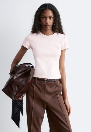 Jeune femme aux cheveux bouclés portant un haut rose pâle à manches courtes et un pantalon marron, tenant un grand sac en cuir marron foncé avec une écharpe noire.