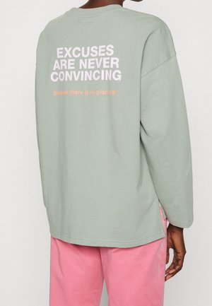 Personne portant une chemise verte claire à manches longues avec le texte "EXCUSES ARE NEVER CONVINCING" et un pantalon rose, vue de dos.
