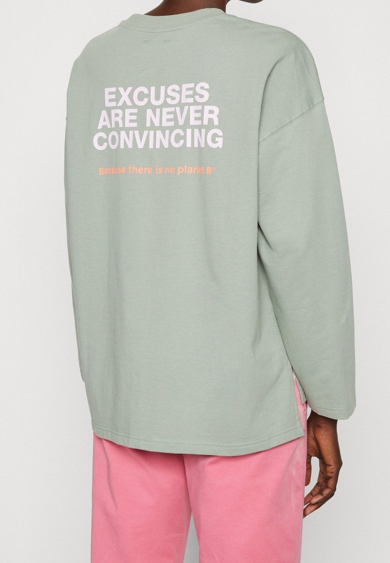 Personne portant une chemise verte claire à manches longues avec le texte "EXCUSES ARE NEVER CONVINCING" et un pantalon rose, vue de dos.