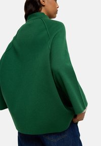 Zielony płaszcz z mieszanki wełny z wysokim kołnierzem, oversize'owymi rękawami i bocznymi rozcięciami. Teksturowana tkanina na białym tle. Casualowy krój.