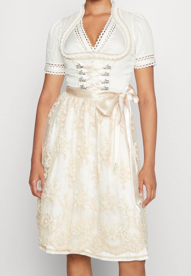 Femme portant une robe traditionnelle blanche et crème avec manches courtes, corset à lacets sur le devant, ruban de satin à la taille et jupe en dentelle florale.