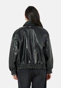 Vrouw gezien van achteren, gekleed in een zwarte jas van imitatieleer met elastische zoom en donkergekleurde, hooggewaste jeans, tegen een witte achtergrond.