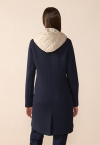 Cappotto blu navy con cappuccio trapuntato beige. Presenta una silhouette elegante, maniche lunghe e dettagli di apertura posteriore. Tessuto dalla texture liscia.