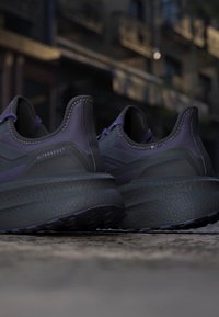 Par sorte og mørkelilla Adidas Ultraboost sneakers placeret på en betonoverflade med en sløret urban baggrund.