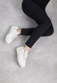 Sneakers bianchi trapuntati con dettagli dorati e un tallone testurizzato, abbinati a leggings neri aderenti, su un pavimento grigio testurizzato.