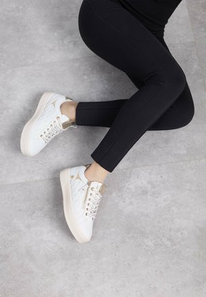 Zapatillas acolchadas blancas con acentos dorados y un talón texturizado, combinadas con leggings negros ajustados, sobre un suelo gris texturizado.