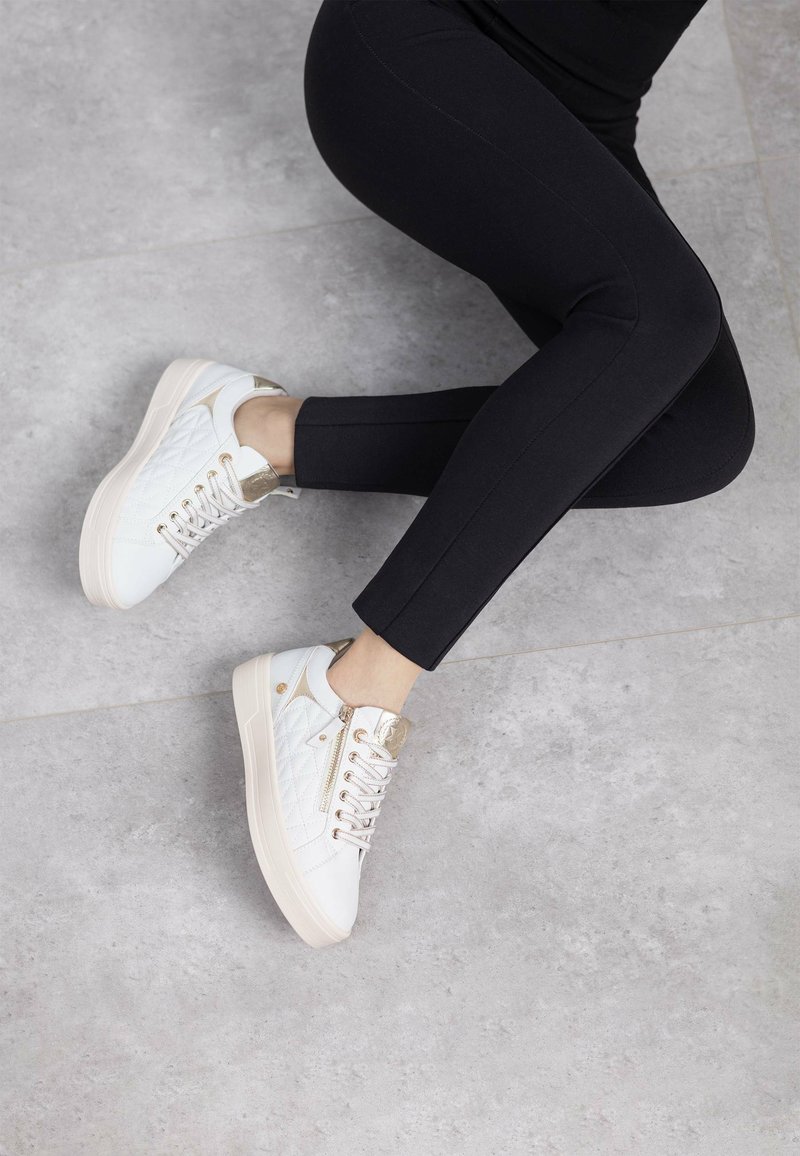 Sneakers bianchi trapuntati con dettagli dorati e un tallone testurizzato, abbinati a leggings neri aderenti, su un pavimento grigio testurizzato.