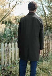 Abrigo negro largo con corte recto, aberturas traseras y un cuello. Combinado con pantalones azul marino, en un fondo natural al aire libre.