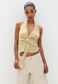 Pletený halter top v svetlej béžovej farbe s detailom uzla vpredu a hlbokým výstrihom. Skombinovaný so svetlom béžovými, voľne strihanými nohavicami.