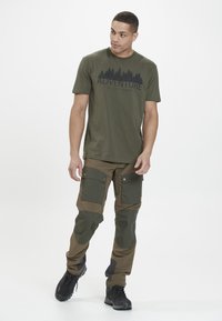 Oliivinvihreä t-paita, jossa on musta "SEIKKAILU ON ULKONA" -grafiikka, sekä tummanvihreät ja ruskeat cargo-housut, joissa on taskuja ja tekstuurisiivouksia.