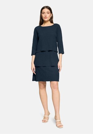 Femme debout portant une tenue en deux pièces texturée sombre avec un haut à manches trois-quarts et une jupe longueur genou, ainsi que des talons ouverts beige.