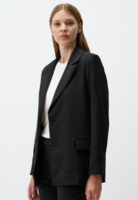 Blazer nero a righe con un design strutturato, revers a punte, due bottoni e tasche frontali. Abbinato a una maglietta bianca sotto.