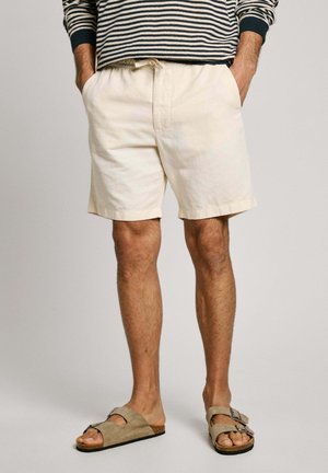Mand iført lyse beige shorts, stribet langærmet skjorte og taupe ruskindssandaler med spænde, med bare ben og hænder i lommerne.