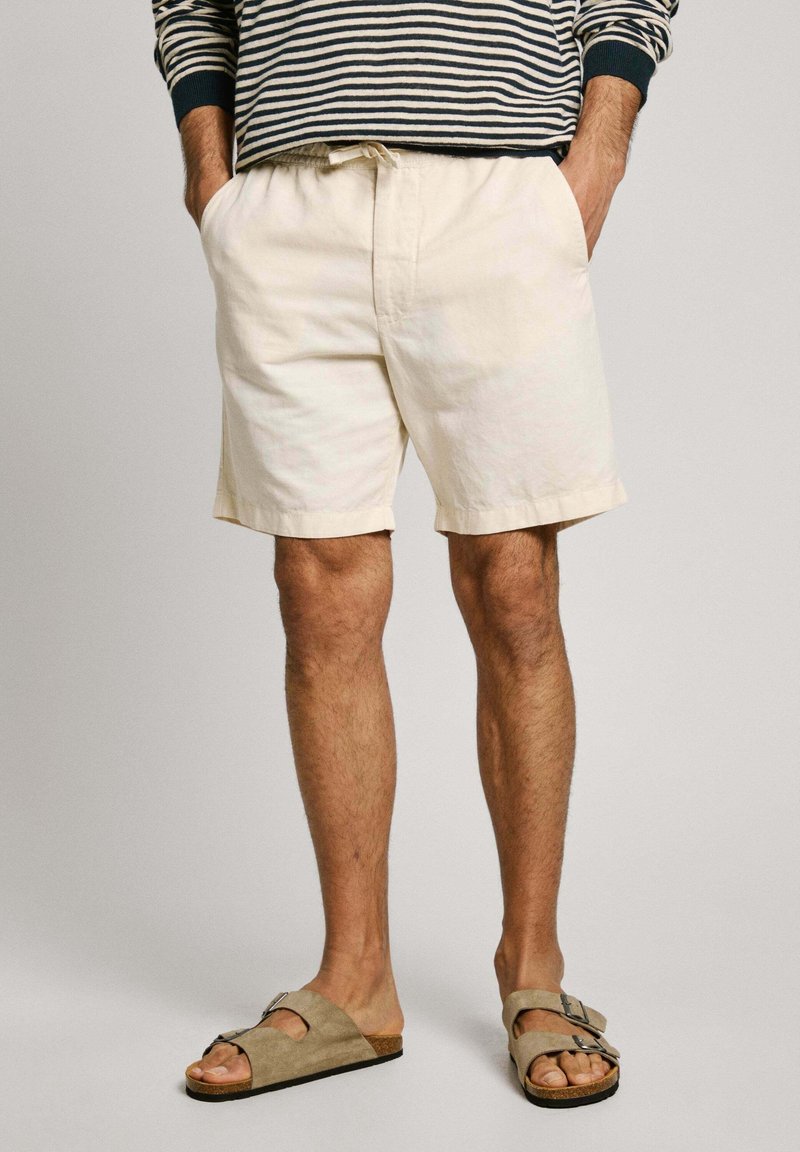 Homme portant un short beige clair, une chemise à manches longues rayée et des sandales en daim taupe à boucle, les jambes nues et les mains dans les poches.