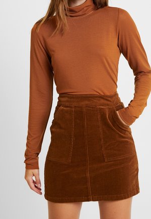 Woman wearing a fitted rust-colored turtleneck and a brown corduroy skirt with front pockets, one hand in pocket and the other relaxed by side.