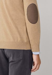 Maglione beige con toppe scure sui gomiti indossato sopra una manica a righe blu e bianche, abbinato a pantaloni grigio scuro.