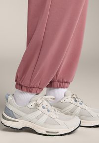 Světlé růžové tepláky s manžetami v kombinaci s bílými a světle modrými sportovními botami, vyrobené z texturované tkaniny a s detaily z různých materiálů.