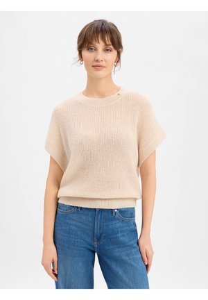Vrouw met lichtbruin haar, gekleed in een beige gebreide trui met korte mouwen en een blauwe jeans met hoge taille, tegen een effen witte achtergrond.