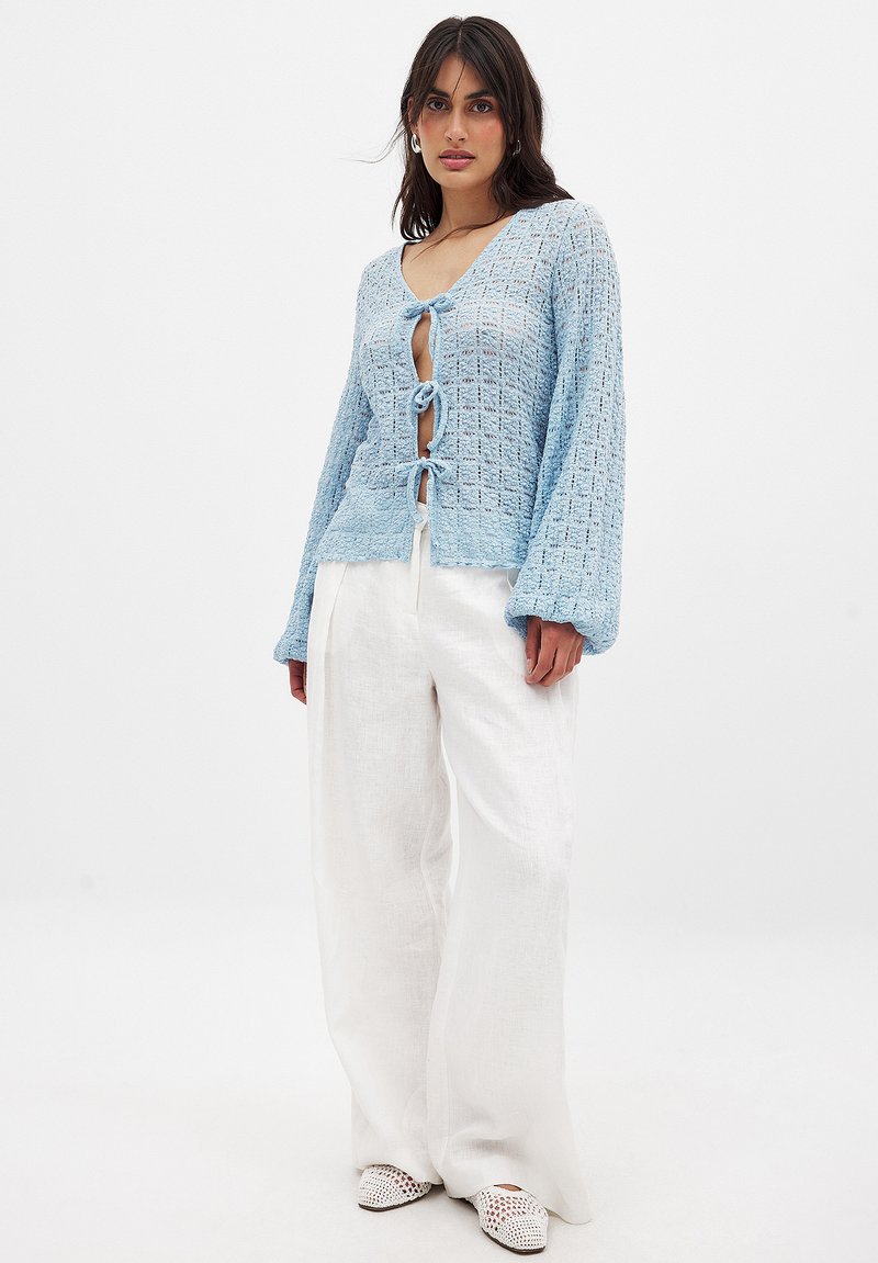 Lichtblauwe gebreide cardigan met een losse pasvorm, voorzien van een V-hals en een sluiting met een strik aan de voorkant. Gepaard met witte wijde broek.