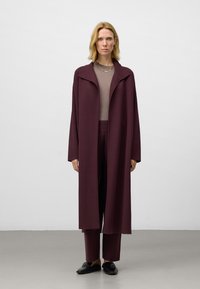 Lang, open voorkant jas in diep bordeaux, gemaakt van zacht materiaal. Daaronder een beige top en bijpassende broek. Eenvoudige zwarte platte schoenen maken de outfit af.