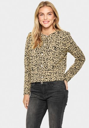 Leopardenmuster, beiger Pullover mit schwarzen Akzenten, langen Ärmeln, Knopfleiste und geripptem Saum, kombiniert mit dunkelgrauen Jeans.