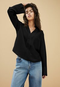 Sort langærmet skjorte med V-udskæring, tekstureret stof, parret med lyseblå højtaljede jeans. Baggrunden er lys beige.