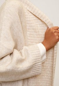 Cardigan léger beige avec des poignets côtelés et un design en tricot texturé. Il présente un col châle et des rayures horizontales subtiles.