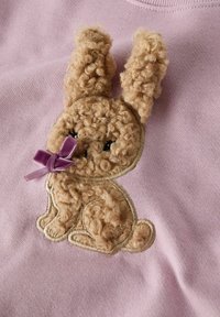 Hele roosa dressipluus, millel on tekstuuriga beež jänkuaplikatsioon, mille karv on lokkis, detailidena mustad silmad ja lilla vibu.