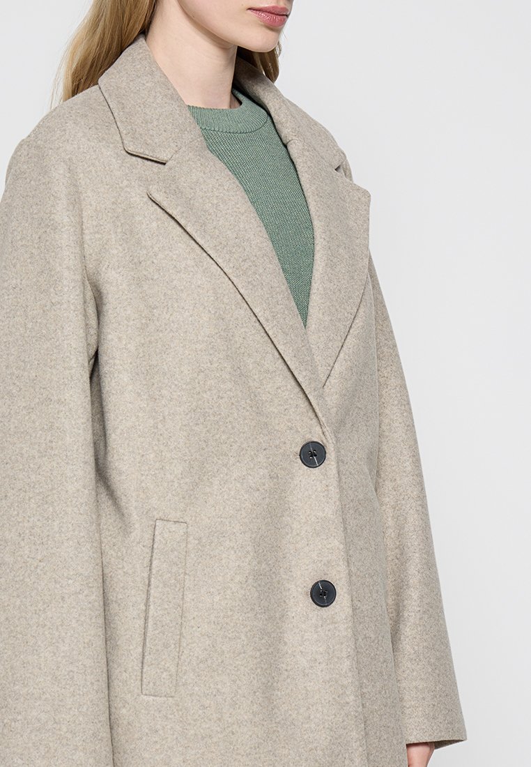 Abrigo de mezcla de lana beige con un cuello entallado, dos bolsillos frontales y botones negros, superpuesto sobre un suéter verde.