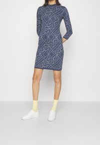 Tmavě modré šaty s geometrickým bílým vzorem, dlouhými rukávy a přiléhavým střihem. Doplněné bílými teniskami a světlými žlutými ponožkami.