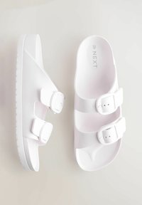 Vita slip-on sandaler med två justerbara spännen och en texturerad sula. Tillverkade av slät, hållbar material med en minimalistisk design.