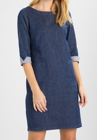 Vestido de mezclilla en azul oscuro, con mangas tres cuartos con puños estampados, dos bolsillos delanteros y un ajuste ligeramente holgado.