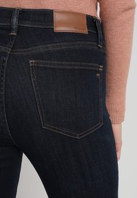 Tmavě modré džínové kalhoty s hladkou texturou, vybavené světlě hnědou koženou nášivkou v pase a zakřivenými zadními kapsami se švy.