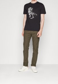 Man som bär svart T-shirt med vit grafisk bild av en mekanisk dinosaurie och texten "BOSS", olivgröna byxor och vita sneakers, står mot vit bakgrund.