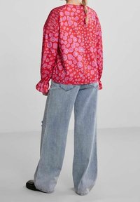 Blusa rossa con motivo circolare rosa, maniche lunghe e polsini arricciati abbinata a jeans azzurri larghi e scarpe nere.