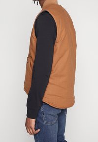 Brązowa bezrękawnik w teksturowanej tkaninie z wysokim kołnierzem. Posiada dwie boczne kieszenie oraz kontrastową czarną koszulę z długim rękawem na dole.