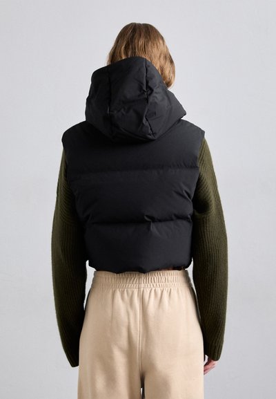 Svart puffer vest med hette, som har en kviltet design. Kombinert med en ribbet grønn genser og beige bukser med elastisk midje.