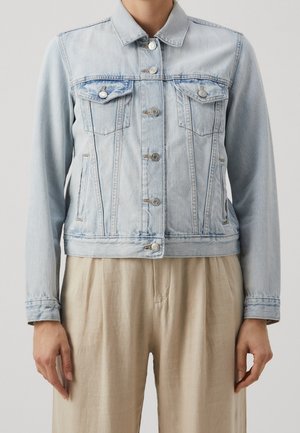 Veste en jean bleu clair avec fermeture à boutons et poches poitrine portée sur un pantalon beige, montrée du cou aux hanches.