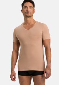 Beżowa koszulka z krótkim rękawem w kształcie litery V, o dopasowanym kroju, wykonana z gładkiego materiału. Połączona z czarnymi dopasowanymi szortami, widocznymi na dolnej krawędzi.